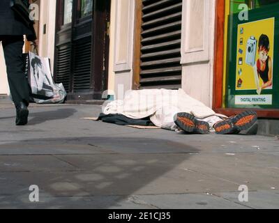 Senza casa travaso destitute addormentato sulla strada vicino allo Strand, con shopper che passa vicino e pubblicità per la stampa digitale in background. I ricchi poveri dividono le disuguaglianze, Foto Stock