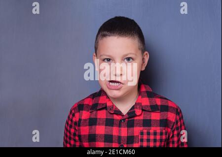 Allegro adolescente europeo con capelli marrone chiaro e un la camicia rossa in plaid crea un volto divertente su un grigio isolato sfondo Foto Stock