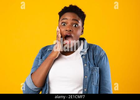 Shocked Plus-Size African Woman guardando fotocamera in posa, sfondo giallo Foto Stock