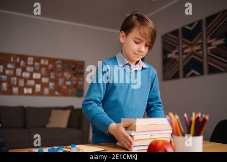 Formazione online a distanza, apprendimento su Internet. Ragazzo di primo grado che studia a casa, tiene un mazzo di libri, preparandosi per la lezione online Foto Stock
