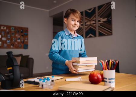 Formazione online a distanza, apprendimento su Internet. Ragazzo di primo grado che studia a casa, tiene un mazzo di libri, preparandosi per la lezione online Foto Stock