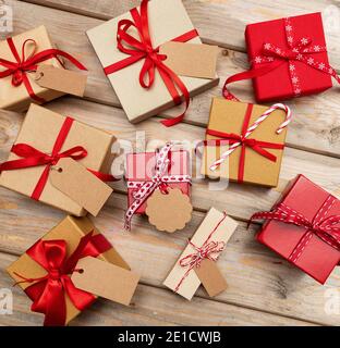 Scatole regalo di Natale con etichette vuote su sfondo di legno. Posa piatta di confezioni attuali fatte a mano, carta da imballaggio kraft, nastro rosso. Ideale per xmas cel Foto Stock