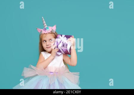 Carina bambina in elegante abito tulle e archetto unicorno celebra la festa di compleanno su sfondo colorato. Faccia divertente con scatola del regalo. Foto Stock