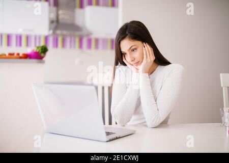 Focalizzato giovane bella donna sta leggendo attentamente roba sul suo laptop mentre si siede al tavolo in una stanza molto luminosa. Foto Stock