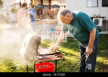 L'uomo felice anziano sta controllando la sua carne su un barbecue mentre la sua famiglia sta godendo in un cortile Foto Stock