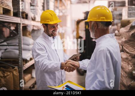 Il lavoro è fatto. Due maschi sorridenti soddisfatti che scuotono le mani dopo aver chiuso un affare di successo. In piedi in grande magazzino vestito di camici bianchi Foto Stock