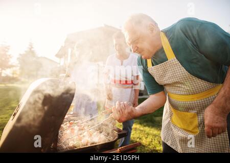 L'uomo felice anziano sta controllando la sua carne su un barbecue mentre la sua famiglia sta godendo in un cortile Foto Stock