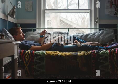 Un giovane uomo si stende sul letto che accarezza il cane nel suo giro e leggere un libro Foto Stock