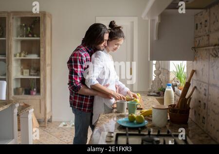 Vista laterale del ragazzo che abbraccia la donna tagliando le mele durante la cottura colazione a casa Foto Stock