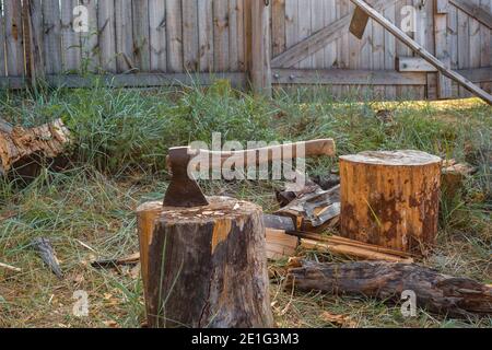 Una grande vecchia ascia si stende da un ceppo di legno su uno sfondo di erba verde e una recinzione in una giornata estiva soleggiata. Un'ascia per tritare il legno. Foto Stock