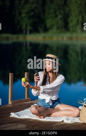 Bella donna felice mangia una mela e facendo selfie mentre si siede su un molo di legno vicino al lago. Foto Stock