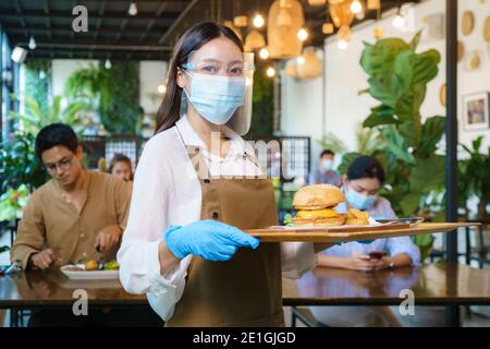 Ritratto attraente cameriera asiatica indossare maschera viso e visiera di protezione tenendo vassoio cibo per servire il pasto al cliente con il custode in background. Nuovo normale