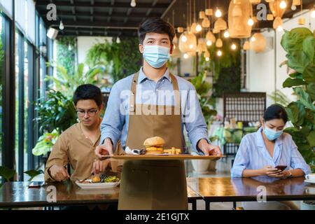 Ritratto attraente cameriera asiatica indossare maschera viso e visiera di protezione tenendo vassoio cibo per servire il pasto al cliente con il custode in background. Nuovo normale