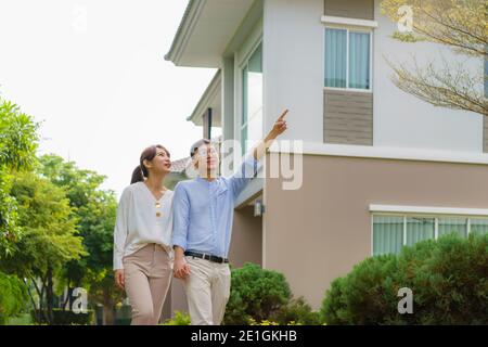 Ritratto di coppia asiatica che cammina e si aggirano insieme guardando felice di fronte alla loro nuova casa per iniziare la nuova vita. Famiglia, età, casa, immobiliare e. Foto Stock