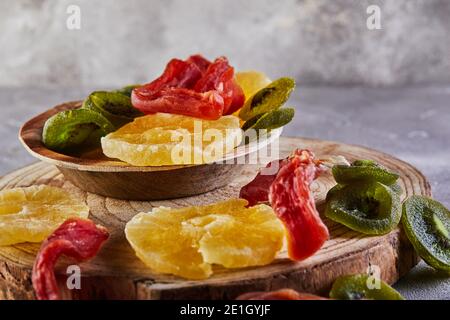 Frutta secca: Anelli di ananas canditi gialli, papaya rossa e kiwi verde su tavola di legno e in un piatto di legno Foto Stock