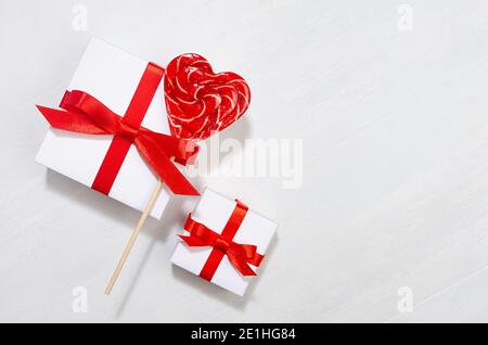 Luminoso sfondo festivo di nozze - scatole regalo bianche con arco rosso, cuori di lecca dolce su tavola di legno bianco, bordo. Foto Stock