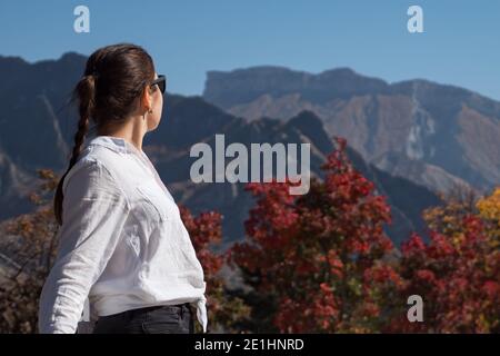 Giovane donna in occhiali da sole ammira le grandi colline rocciose di marrone colori e alberi rossi sotto il cielo blu chiaro in autunno vista giorno sul retro Foto Stock