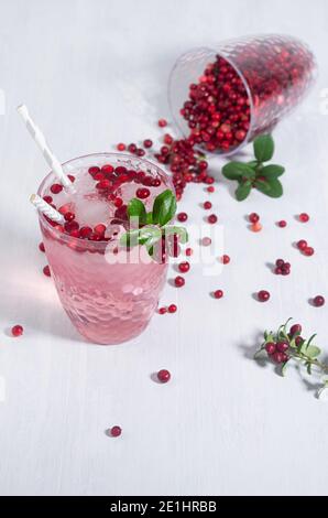 Cocktail rosa con mirtillo, ghiaccio e foglie verdi sparse in fascio di sole su tavolo di legno bianco, verticale. Foto Stock