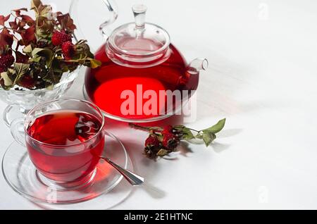 Colazione con tè biologico rosso biancospino in teiera trasparente, tazza, frutti di bosco biancospino, rosa anca alla luce del sole con ombra su tavola di legno bianco, guardaroba. Foto Stock