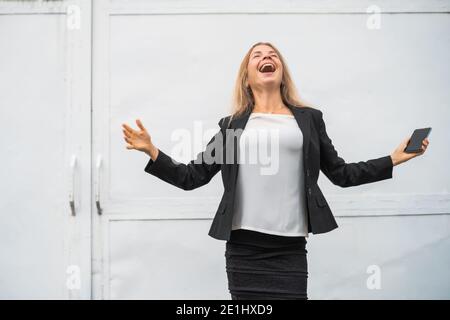 Ritratto esterno di una donna d'affari moderna e estatica che è gioiosa. Ha uno smartphone. Foto Stock