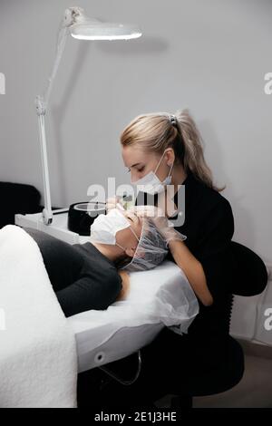 Il padrone permanente del trucco fa la procedura di rimozione dei capelli facciali da cera calda in salone, preparazione prima del tatuaggio permanente del trucco sulle sopracciglia del cliente. Sicuro Foto Stock