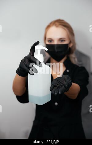 Giovane donna offuscata in nero e guanti e maschera chirurgica con alcool blu nelle mani in bottiglia a fuoco. Utilizzo di gel igienizzante per le mani per la pulizia Foto Stock