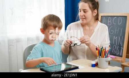 Madre che mette gli occhiali sul suo figlio astuto che gioca sul computer del tablet e che fa i compiti. Problema del bambino con gli occhi e la vista. Foto Stock