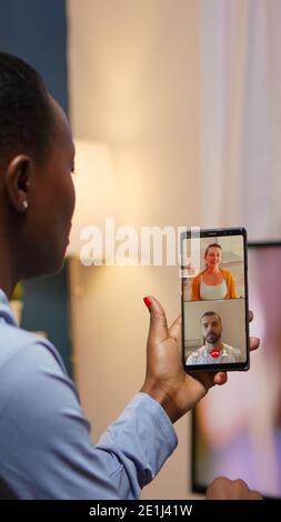 Donna nera imprenditore remoto che ha una videoconferenza con un amico che utilizza uno smartphone seduto in salotto a tarda notte. Dipendente occupato che utilizza una rete wireless con tecnologia moderna che fa straordinari per il lavoro Foto Stock