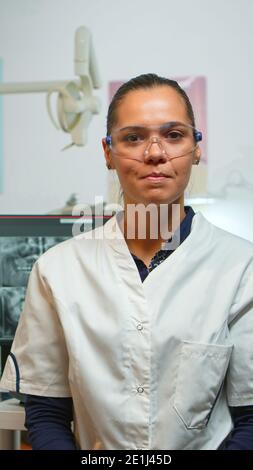 Medico stomatologo che parla su web cam con i pazienti di igiene orale guardando la fotocamera mentre il paziente in attesa di infermiere in background. Assistente dentale che prepara l'uomo per la chirurgia stomatologica. Foto Stock