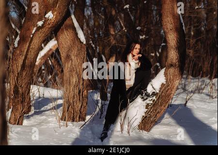 Giovane ragazza appoggiata sull'albero coperto di neve nella foresta d'inverno. Giornata invernale soleggiata Foto Stock