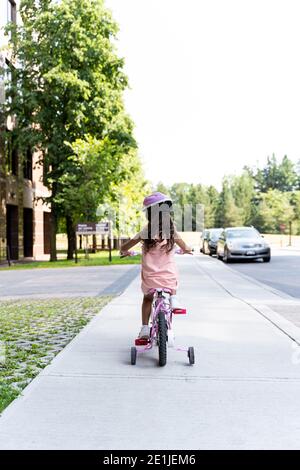 Giovane ragazza in abito rosa e casco in bicicletta rosa sul marciapiede di strada, visto da dietro Foto Stock