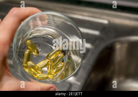 Un vaso di vetro tenuto in mano con capsule di colore oro giallo - integratori alimentari, vitamine, medicinali Foto Stock