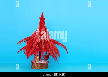 Albero di Natale decorativo rosso in miniatura con piccoli ornamenti posti sfondo blu brillante in studio Foto Stock