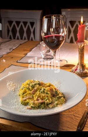 Fettuccine di Carbonara con asparagi. Spaghetti all'uovo con pancetta, uova e parmigiano in piatto su tavolo da trattoria con bicchiere di vino e candela accesa Foto Stock