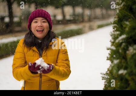 Giovane donna giapponese asiatica felice e attraente in giacca invernale e beanie godendo nevicate al parco cittadino giocando allegro circondato dalla neve durante Foto Stock