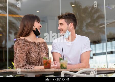 Una giovane coppia indossa maschere facciali che sorridono l'una all'altra in una terrazza all'aperto. Concetto di svago e sicurezza. Foto Stock