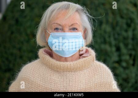 Signora anziana che indossa una maschera protettiva per il viso all'aperto nel giardino in inverno in un ritratto frontale durante il Covid-19 pandemia Foto Stock