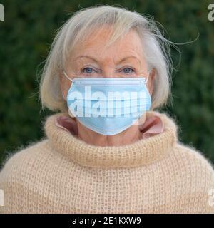 Signora anziana che indossa una maschera protettiva per il viso all'aperto nel giardino in inverno in un ritratto frontale durante il Covid-19 pandemia Foto Stock