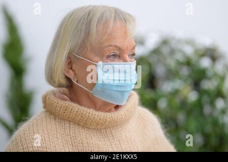 Donna anziana disperata durante la pandemia di Covid-19 che si indossa all'aperto una maschera protettiva monouso che guarda da parte tranquillamente Foto Stock