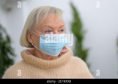 Donna anziana disperata durante la pandemia di Covid-19 che si indossa all'aperto una maschera protettiva monouso che guarda da parte tranquillamente Foto Stock