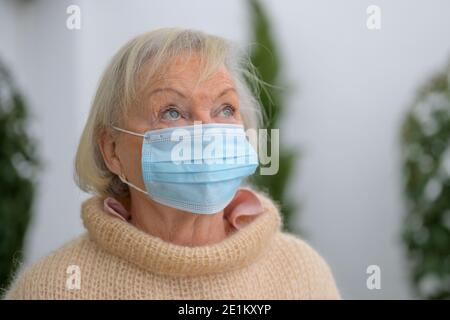 Donna anziana dagli occhi blu che indossa una maschera facciale come protezione durante La pandemia di Covid-19 in piedi all'aperto guardando in su con un attento espressione in a. Foto Stock