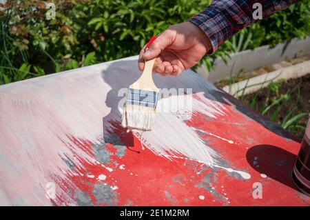 painter dipinge la superficie bianca. L'immagine è uno sfondo bianco appena dipinto e un pennello in mano su un tavolo rosso. Foto Stock