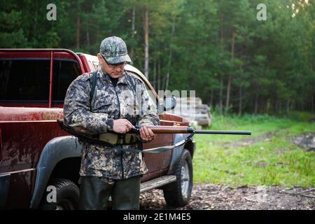 Cacciatore uomo in piedi con fucile da fucile vicino alla sua auto all'aperto preparazione per la caccia agli uccelli nella foresta autunnale Foto Stock