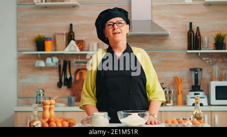 Donna anziana felice che indossa bonete guardando la macchina fotografica nella sala da pranzo a casa. Vecchio panettiere pensionato con uniforme da cucina preparazione ingredienti da forno a tavola pronti per cucinare pane, torte e pasta fatti in casa. Foto Stock