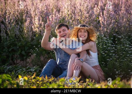 La coppia felice in amore si siede abbracciando da campo di salvia fiorente. Un giovane con bella ragazza in cappello di paglia si divertono, si imbrogliano intorno, agitando il loro ha Foto Stock