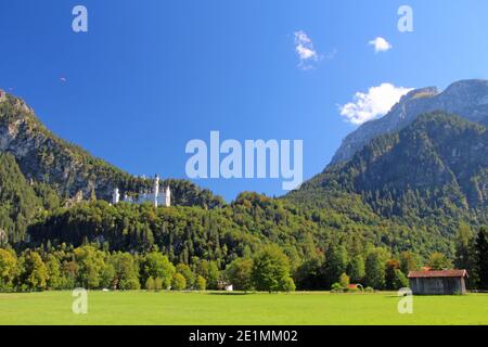 German Fairy tale Castello Neuschwanstein in panoramico e romantico panorama delle alpi con montagne verdi, cielo blu, sole e nuvole Foto Stock