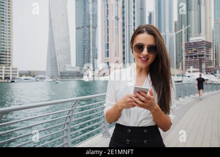 Bella giovane donna d'affari moderna in piedi in una Marina di Dubai e guardando il suo telefono. Foto Stock