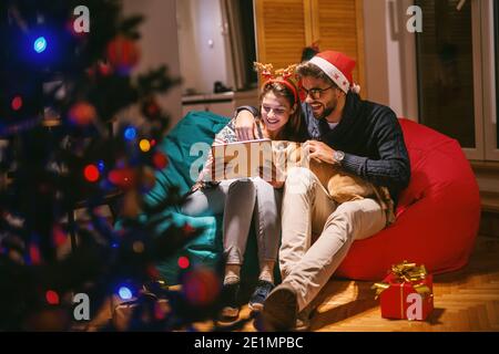 Accoppia usando il tablet mentre sei seduto in salotto. Uomo che tiene il cane. Sul capo cappello di babbo natale e fascia di renna. In primo piano albero di Natale. Natale Foto Stock