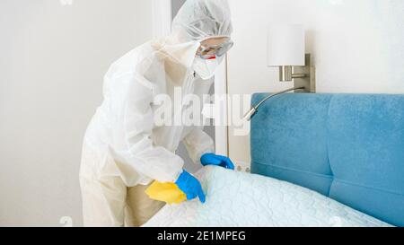 Casalinga in tuta medica protettiva e maschera facendo pulizia e. fare il letto a casa Foto Stock