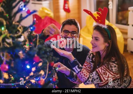 Coppia che decorano l'albero di Natale. Sul capo cappello di babbo natale e fascia di renna. Interni domestici, concetto di vacanze di Natale. Foto Stock